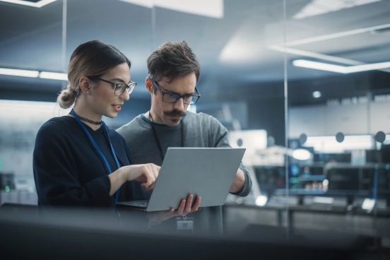 Une photographie d’un homme et d’une femme debout côte à côte en train de regarder un écran d’ordinateur portable dans un environnement de bureau