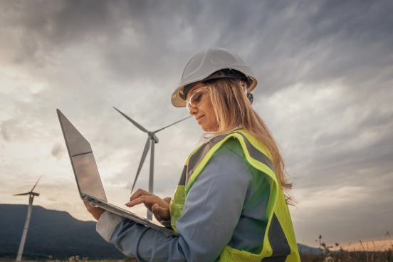 Une photo d’une personne portant un haut haute visibilité et un casque de sécurité devant une éolienne faisant fonctionner un ordinateur portable