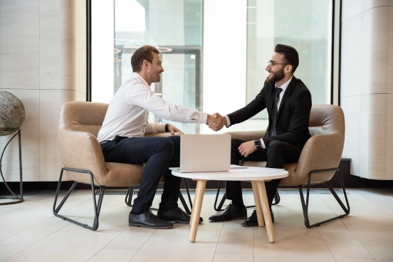 Une photo de deux hommes souriants en tenue d’affaires formelle assis à une table se tendant la main pour se serrer la main